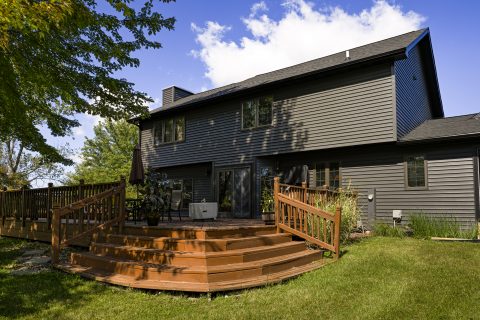 Contemporary gray house with wooden deck and lush green yard.