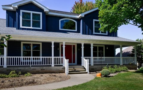 Modern blue house with white porch railing and lush green yard.