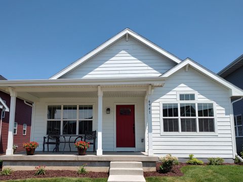 White house with red front door and large windows, front porch with benches and flower pots.