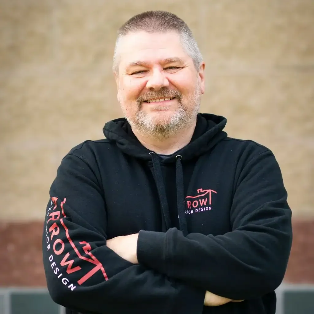 Smiling man in black hoodie with Arrow Exteriors logo, arms crossed, outdoors.